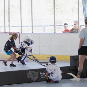 Youth participants compete in a ball hockey game on an indoor rink. One player wearing goalie equipment defends the net while other participants and coaches watch from the sidelines.