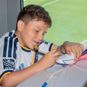 A young participant in a LA Galaxy jersey uses a gold marker to decorate a soccer cleat at a craft station overlooking the stadium field.