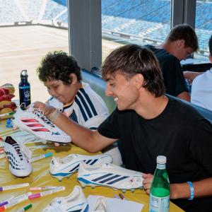 A LA Galaxy player Julian Aude and a young participant sit at a table covered with soccer cleats and colorful markers, decorating footwear during a community event overlooking the stadium field.