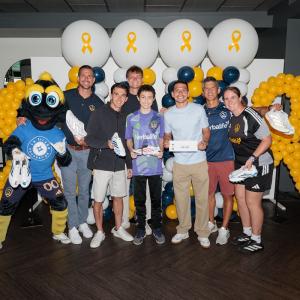 LA Galaxy front office staff and youth participants and mascot Cozmo pose with decorated soccer cleats in front of blue, white, and gold balloon displays featuring gold ribbon symbols.