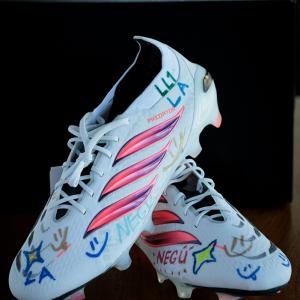 A pair of white Adidas soccer cleats decorated with colorful drawings, stars, smiles, and inspirational messages are displayed on a table after a customization activity.