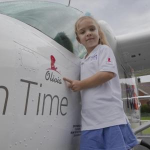 Young girl standing next to plane, pointing at her name (Olivia) printed on it.