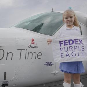 Olivia standing next to the plane with her name on it holding a "FedEx Purple Eagle" sign.