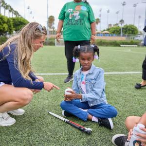 Ashley Sessa kneels beside a seated student holding a field hockey ball, with a stick resting on the turf during a demonstration.