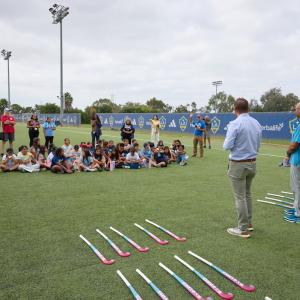 Students sit together on the field listening to two speakers, with rows of colorful field hockey sticks arranged in the foreground.