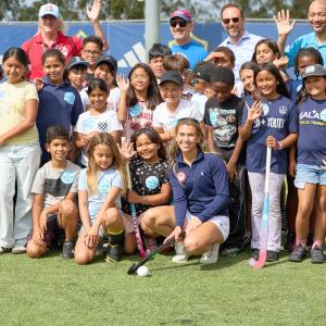 Students and event representatives gather for a group photo on the field, holding field hockey sticks and waving toward the camera.