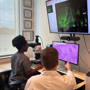 A person looks through a microscope while a person in a white coat points to a screen