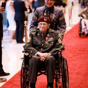 A person pushing a war veteran in a wheelchair down a red carpet 