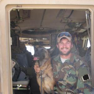 A smiling person in camoflage, sitting in an open vehicle door with a dog