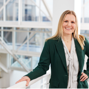 Maureen McHugh smiles at the camera and stands holding a railing at the top of a set of white steps at Bath & Body Works’ home office in Columbus, Ohio. Sun shines through a wall of windows in the background.