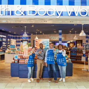 Four Bath & Body Works associates stand at the entrance of a store smiling and giving a “number 1” hand signal to the camera. A colorful selection of candles, hand soaps, lotions and other products are displayed on tables behind them.