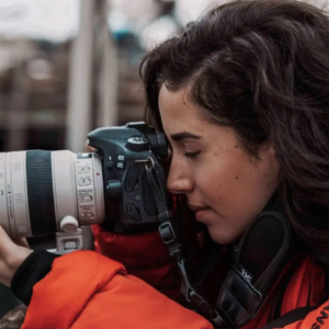 Brunette woman in orange jacket holding a camera