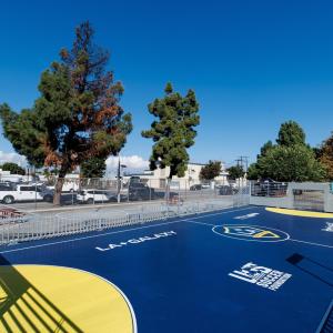 Wide view of a newly installed LA Galaxy-branded mini-pitch with blue and yellow court markings, surrounded by fencing and nearby trees.