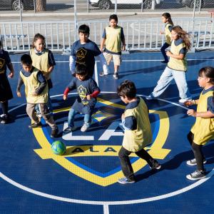 Children wearing LA Galaxy jerseys and pinnies play soccer around a ball on a branded mini-pitch court.
