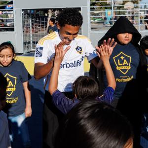 LA Galaxy player in white jersey high-fives youth participants wearing LA Galaxy shirts on a small outdoor soccer court.