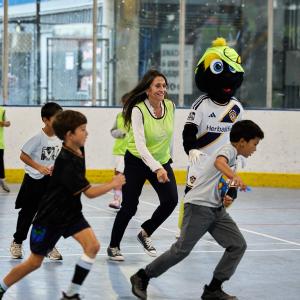 Children run across an indoor court alongside an adult wearing an LA Galaxy training vest and the LA Galaxy mascot.