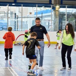A child dribbles a soccer ball on an indoor court as adults in LA Galaxy apparel and other children participate nearby.