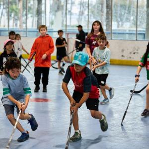 Children play ball hockey on an indoor court, using hockey sticks to compete for an orange ball.