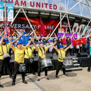 Groups of people in matching colored shirts playing instruments in a stage outside a stadium "Westham United"