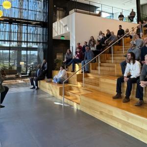 An audience of people sat on a staircase, listening to 3 speakers 