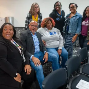 Three people sat next to each other on chairs with four other people stood behind them