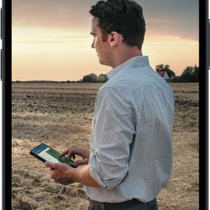 A mobile phone with a photo on the screen on a person stood in a field holding a tablet PC