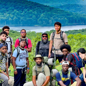 A group in hiking gear, posing for the camera with a tree-covered hill and a lake behind them