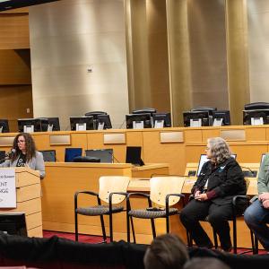 Two people in chairs listening to a speaker behind a podium