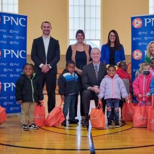 Adults and children posing in a gymnasium next to PNC banners, with orange bags