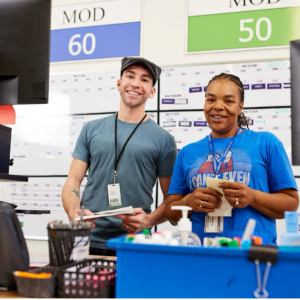 Two Bath & Body Works associates stand smiling behind a table with computer monitors and office supplies at a Bath & Body Works distribution center in Columbus, Ohio. A large calendar is posted behind them.