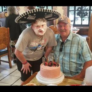 A person in a large sombrero with cream on their face, posing behind a cake with another person sitting at a table