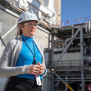 Nicole Flipping stnading outside wearing a hard hat and safety glasses. Industrial pipes and scaffolding behind her.