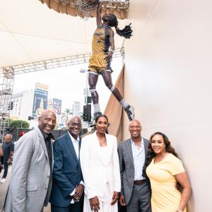 Group poses together beneath a bronze statue depicting Los Angeles Sparks basketball player Lisa Leslie extending toward the basket.
