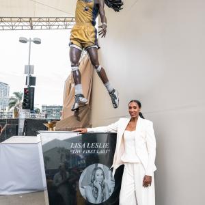 Lisa Leslie poses beside a plaque and a bronze statue depicting a her as basketball player dunking a basketball in a hoop.
