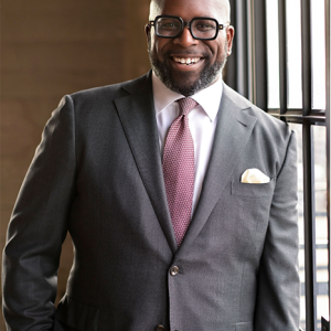 Maurice Cooper is pictured in a grey suit leaning against a sunny window and smiling directly at the camera.