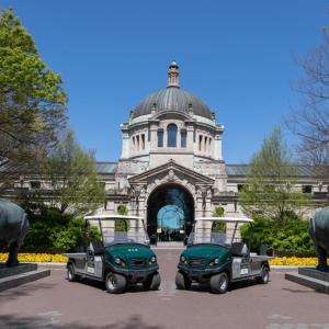 Two electric utility vehicles are parked in front of Bronx Zoo's Central Building on a sunny day. Flanking the vehicles are two animal statues.