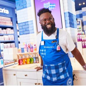 A male Bath & Body Works associate stands in store smiling at the camera in a blue apron with Bath & Body Works products in the background. He leans against a sink where multicolored hand soap bottles are displayed.