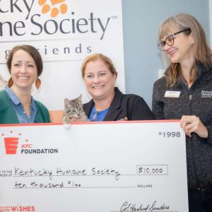 Three people holding an oversized check, with a cat peeking over the top