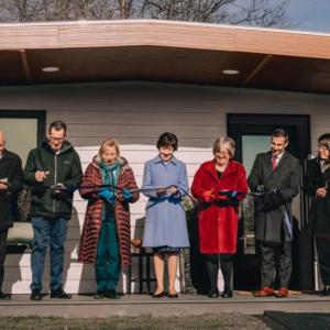 A group of people standing on the porch of the 3D printed home each cutting a portion of the ribbon.