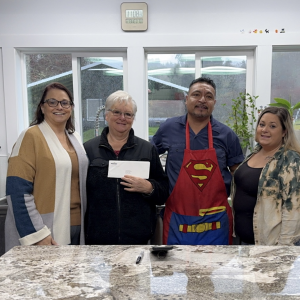 Four people standing together behind a marble table, one holding a check and one in a Superman apron