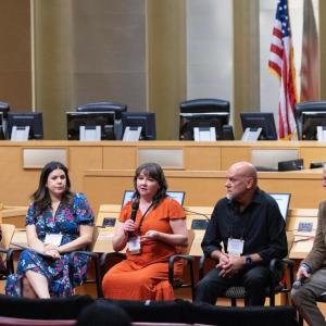 Five people seated, one in a red dress speaking into a microphone