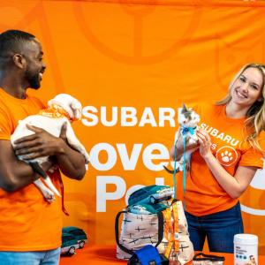 Two people holding a puppy and a kitten, stood in front of an orange Subaru Loves Pets backdrop 