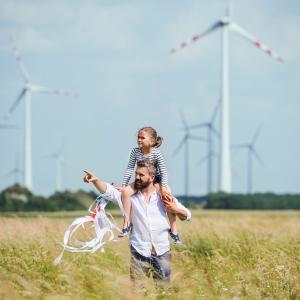 An adult with a child on their shoulders pointing into the distance. Wind turbines in a field behind them. 
