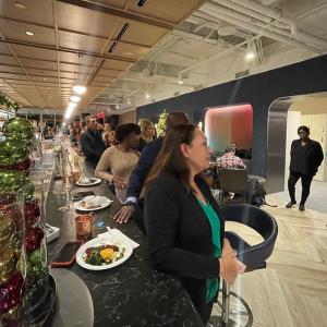 A group of people eating at a long bar, standing and turned to face the opposite side of the room.