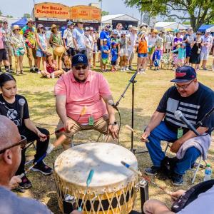 A group sitting around and beating a drum as others watch.