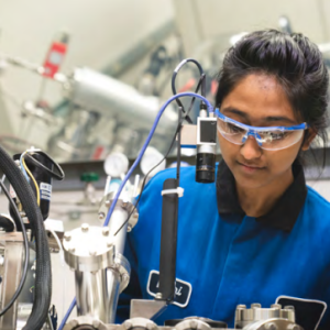 A person in blue and black uniform wearing safety glasses in a lab setting.