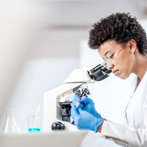 Person wearing safety goggles and a lab coat writing on a test tube next to a microscope