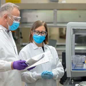 Two scientists look over paperwork. They are dressed in lab coats and personal protection equipment.