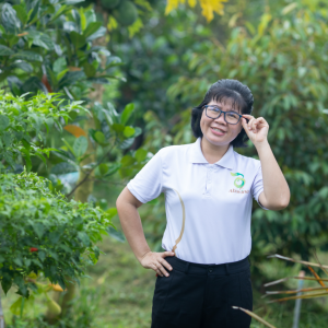 Nguyen Thi Kim Thoa standing in an orchard, touching the side of her glasses. Wearing an Abavina logo shirt