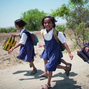 four smiling children running on a dirt road, carrying school bags